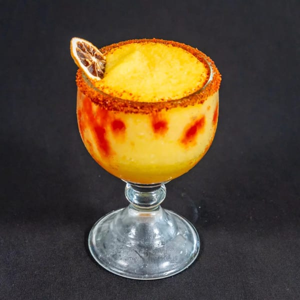 A vibrant yellow frozen drink in a stemmed glass, garnished with a dried orange slice and chili rim.