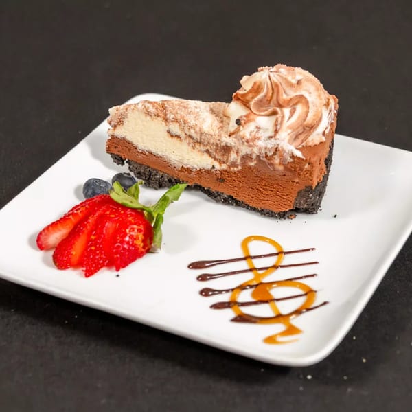 A slice of chocolate and vanilla cheesecake with whipped cream, strawberries, blueberries, and caramel drizzle.